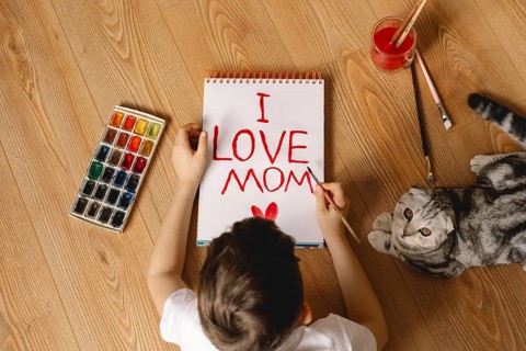 A child paints I LOVE MOM on a notebook using red watercolors, while a curious cat lies beside them