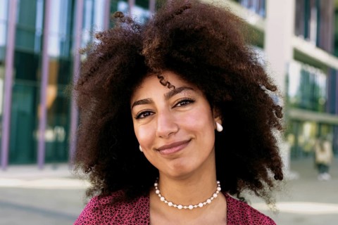 A young Arab woman with a radiant smile and afro hair poses outdoors looking at camera. She wears a