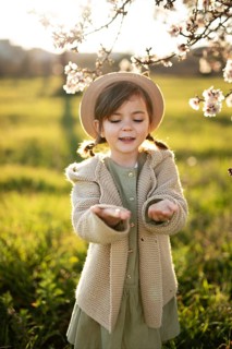 A joyful little girl with a hat stands under almond blossoms, smiling as she extends her hands to