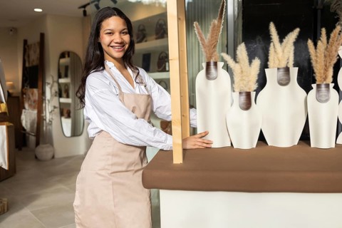 A woman wearing an apron arranges decorative vases on a counter in a chic bag store. Shelves with