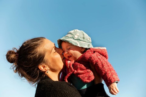 A mother wearing a dark sweater kisses her baby, dressed in a red sweater and green hat, against a