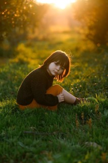 A young child sits on vibrant green grass with orange trees, bathed in the warm, golden glow of a