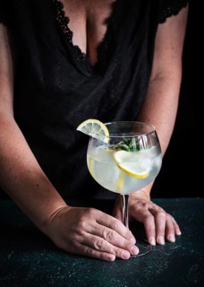 A woman wearing a black dress holds a refreshing gin tonic garnished with lemon slices and mint.
