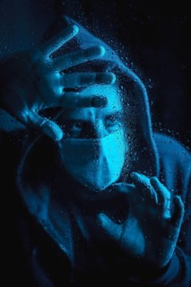 A young caucasian man with a mask looking out the window in the covid19 quarantine, with blue