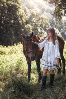 A young girl in a white dress gracefully pets a brown horse in a scenic sunlit field. The ambiance