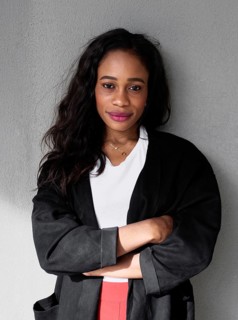 A confident black woman stands with arms folded, wearing a black jacket and white top. Her
