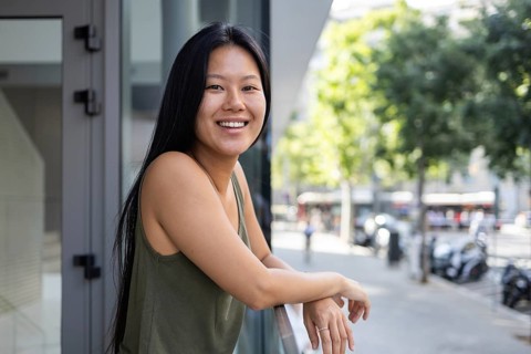 A Chinese woman smiles while leaning on a balcony in a bustling city. The sun shines brightly,