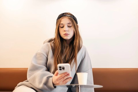 A teenage girl in a casual hoodie and cap is intently using her smartphone while sitting on a