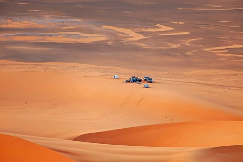 Camp with cars and tents in a vast, tranquil sandy desert landscape under an orange-coloured sky,