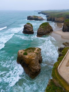 Waves crashing against large, grass-covered rocks along a steep, rugged coastline, aerial view,