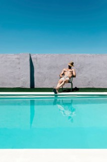A man takes a nap relaxing on a chair by a swimming pool under a clear blue sky. He wears a straw