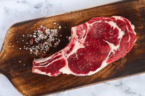 Raw rib eye steak, cowboy steak, wooden chopping board, marble table, top view, no people