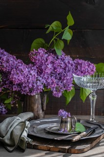 An elegant table setting with vibrant lilac blooms in a rustic pitcher. Vintage silverware and