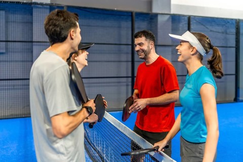Four friends laugh and chat on an indoor pickleball court, holding paddles after a game, enjoying