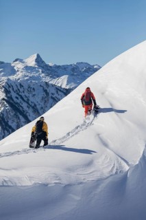 Two snowboarder friends trek up a snowy mountain slope under a clear blue sky. They carry