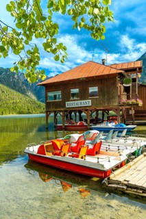 Colorful pedal boats sit docked below a wooden restaurant on lake dobbiaco with the dolomites
