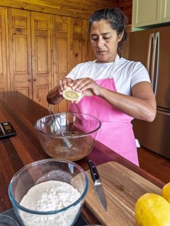 A woman in a pink apron squeezes a lemon into a large glass mixing bowl. Flour and kitchen utensils