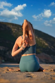 A woman sits in Gomukhasana, stretching in the peaceful embrace of a beach as the sun sets behind