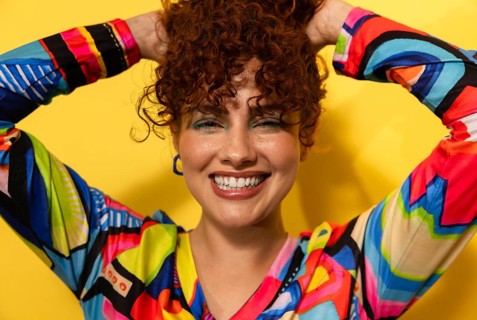 A joyful woman with curly hair smiles brightly, her hands playfully lifting her vibrant, colorful