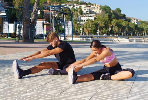 A woman and man engage in stretching exercises on a sunny seaside promenade, surrounded by palm
