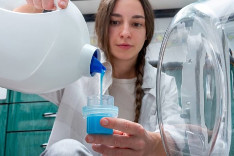A young woman carefully pours bright blue liquid detergent into a cap while sitting beside a