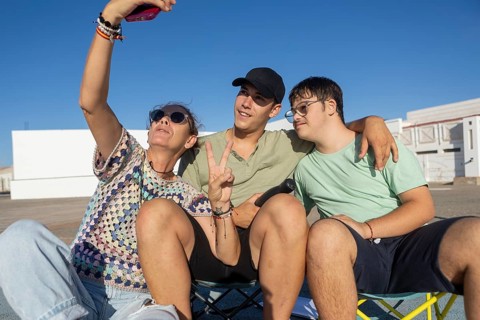 A Down syndrome teenager sits with family members outdoors, laughing and taking a selfie The scene