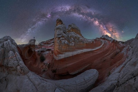 A man stands atop rock formations in White Pocket, Arizona, USA. The night sky is illuminated by