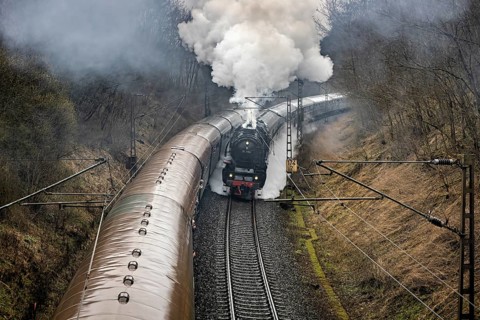 Steaming train passes through foggy landscape, surrounded by trees, on winding tracks, historic