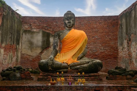 A Buddha statue adorned with an orange robe sits in the midst of ancient brick ruins of Ayutthaya.
