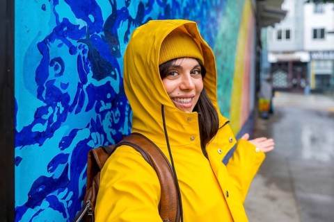 Young woman wearing a yellow raincoat and beanie smiling while standing by a colorful street art