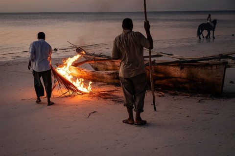 Two local men use a small fire on a sandy beach at twilight to repair or seal a traditional wooden