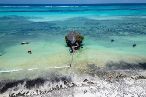 Aerial view of the building surrounded by turquoise water and white sand beach with boats under a