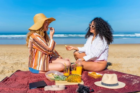 Two diverse women sitting on a blanket on the sand, sharing snacks and orange juice, enjoying a