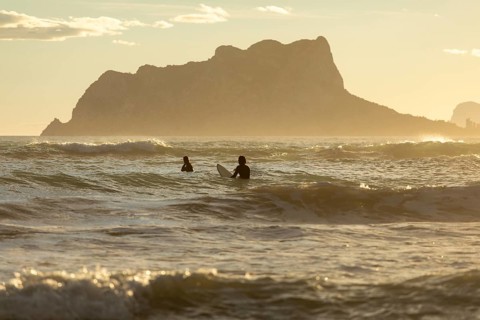 Two surfers enjoy the tranquil waves of the Mediterranean Sea at sunrise, with the Costa Blanca