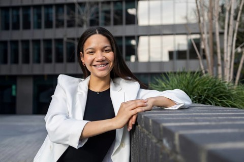 A confident businesswoman in a white blazer and black top smiles outside a modern office building.