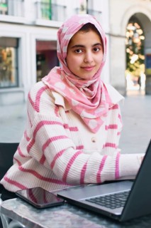 A arab student woman in a pink hijab and striped sweater uses a laptop at an outdoor cafe. Her