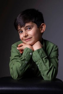 Young boy with dark hair and a content expression rests his chin on his hands, wearing a green