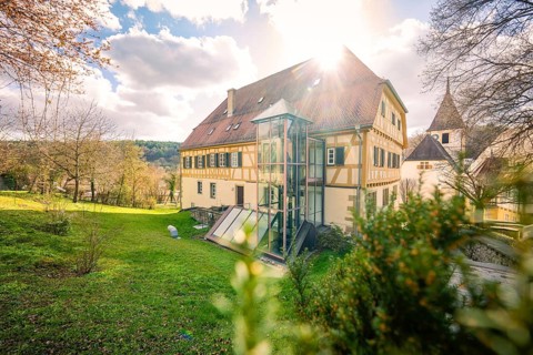 Timbered house with sunshine in the background, surrounded by a green garden and trees, Deufringen