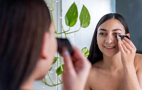 A woman smiles while applying makeup in front of a mirror in makeup session. A green plant with