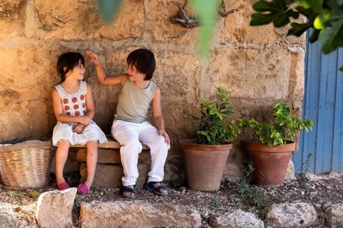 Two children sit on a stone bench near a rustic wall, with potted plants and a woven basket nearby.