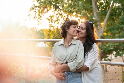 A smiling lesbian couple embraces warmly against a railing at a park. Sunlight filters through the