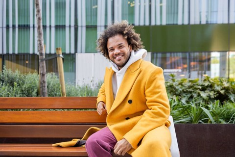 Afro man with curly hair in a bright yellow coat and pink pants sits on a wooden bench, smiling