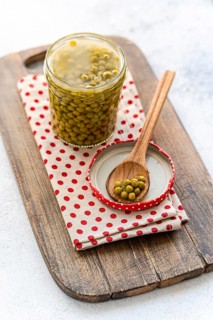 A jar of green homemade organic peas sits on a rustic wooden board, accompanied by a wooden spoon
