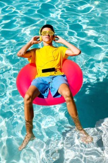 A teenage boy wearing a yellow shirt and sunglasses enjoys music while floating on a pink