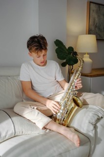 A young boy in casual clothing plays a saxophone while sitting on a sofa. The natural light
