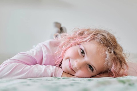 A young child with curly hair lies on a bed, smiling gently. The child wears a light sweater,