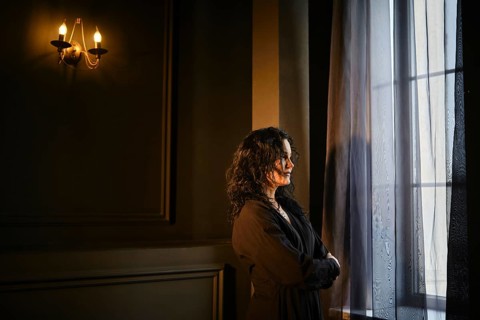 A woman stands by a window in a dark room with soft light from a wall lamp. She gazes outside, lost
