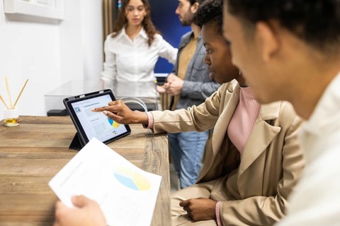 A team gathers around a tablet, focusing on data and charts during a workplace discussion. The