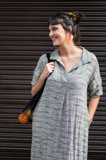 A joyful woman in a casual grey dress stands outdoors, smiling, with colorful earrings and a