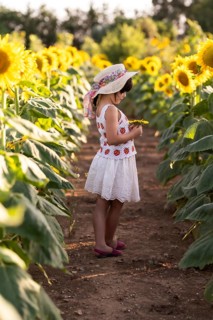 A young girl in a summer dress and sun hat stands in a vibrant sunflower field, holding a sunflower